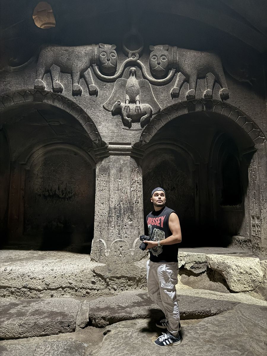Moi debout dans une salle du monastère de Geghard entièrement creusée dans la roche, sous un relief sculpté montrant deux lions encadrant un aigle qui tient un agneau dans ses serres