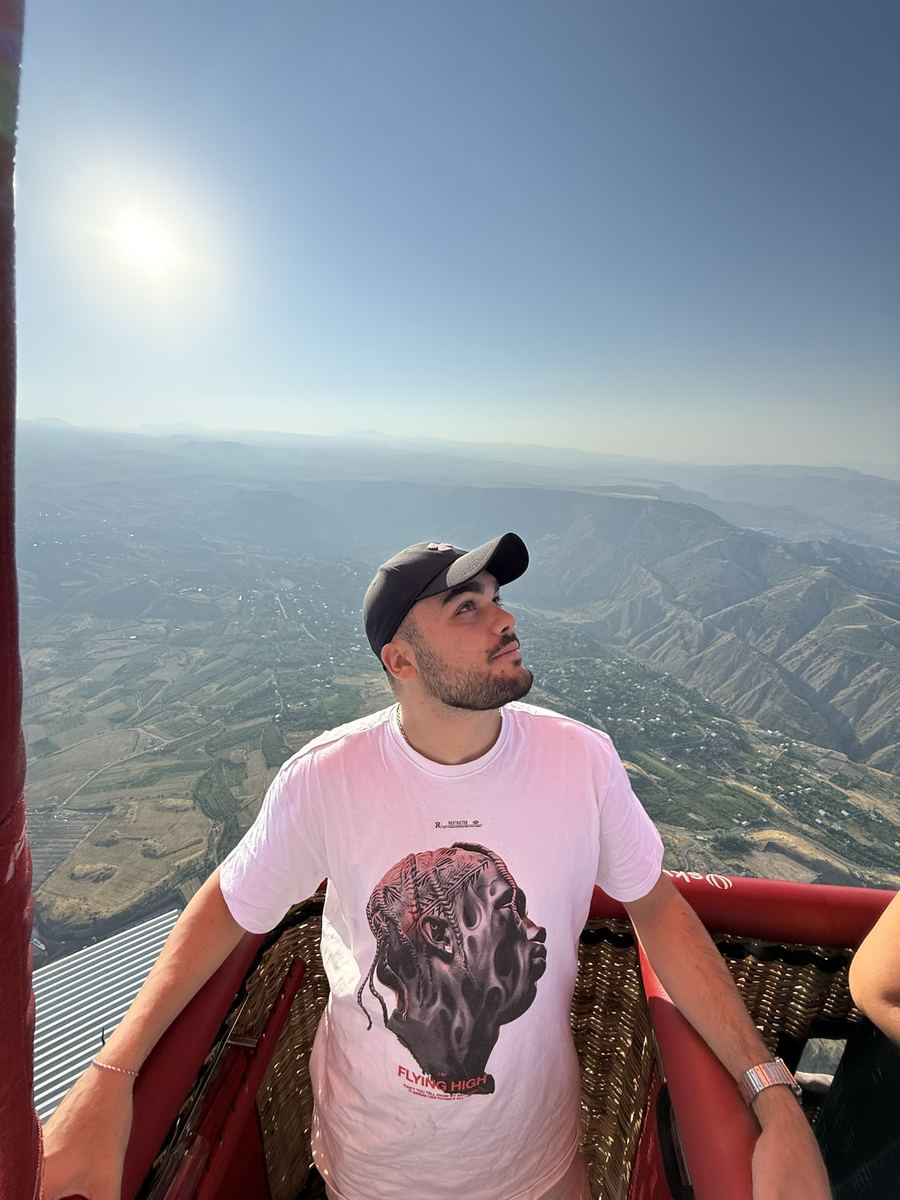 Moi en casquette et t-shirt blanc, accoudé au bord d'une nacelle de montgolfière rouge, une vallée de montagnes et de villages s'étendant très loin en contrebas sous un soleil haut
