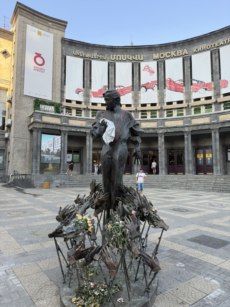 La statue en bronze de Charles Aznavour, bras tendus dans la position du chant, dressée sur un socle en forme de mains sculptées couvert de bouquets de fleurs, devant la façade à colonnes du cinéma Moscou à Erevan