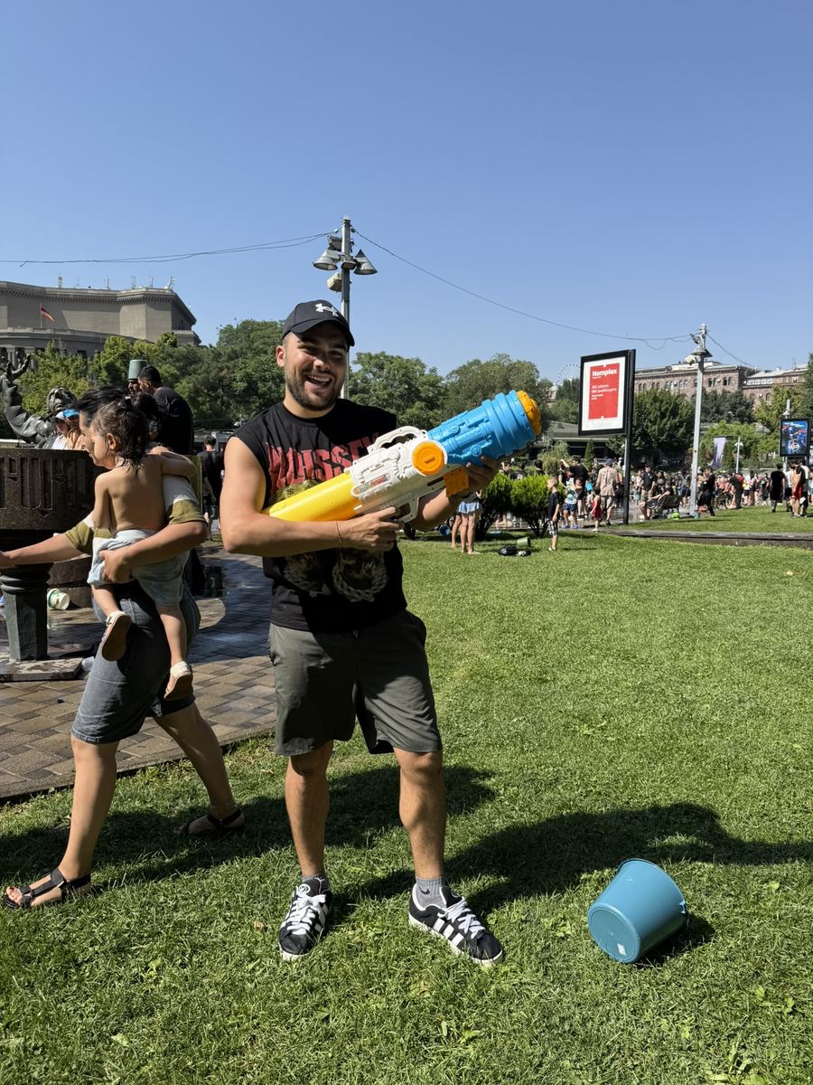 Moi en casquette et débardeur, un gros pistolet à eau jaune et bleu dans les bras, sur une pelouse d'Erevan au milieu d'une foule trempée, un seau bleu renversé au sol