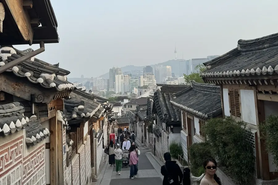 La ruelle en pente de Bukchon bordée des toits de tuiles grises des hanok, avec les tours du centre de Séoul et la N Seoul Tower dans la brume au fond