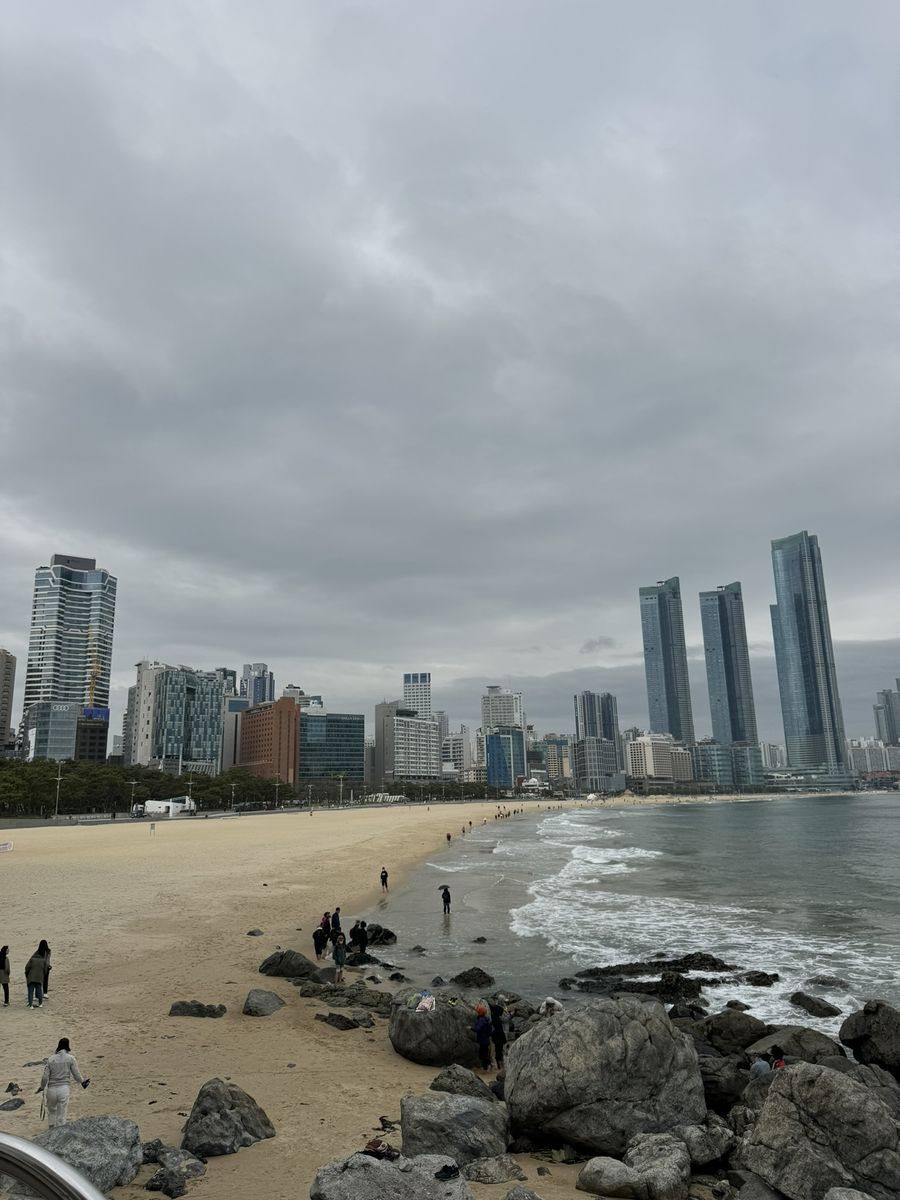 La plage de Haeundae à Busan sous un ciel gris : sable clair, rochers au premier plan, rangée de gratte-ciel en fond