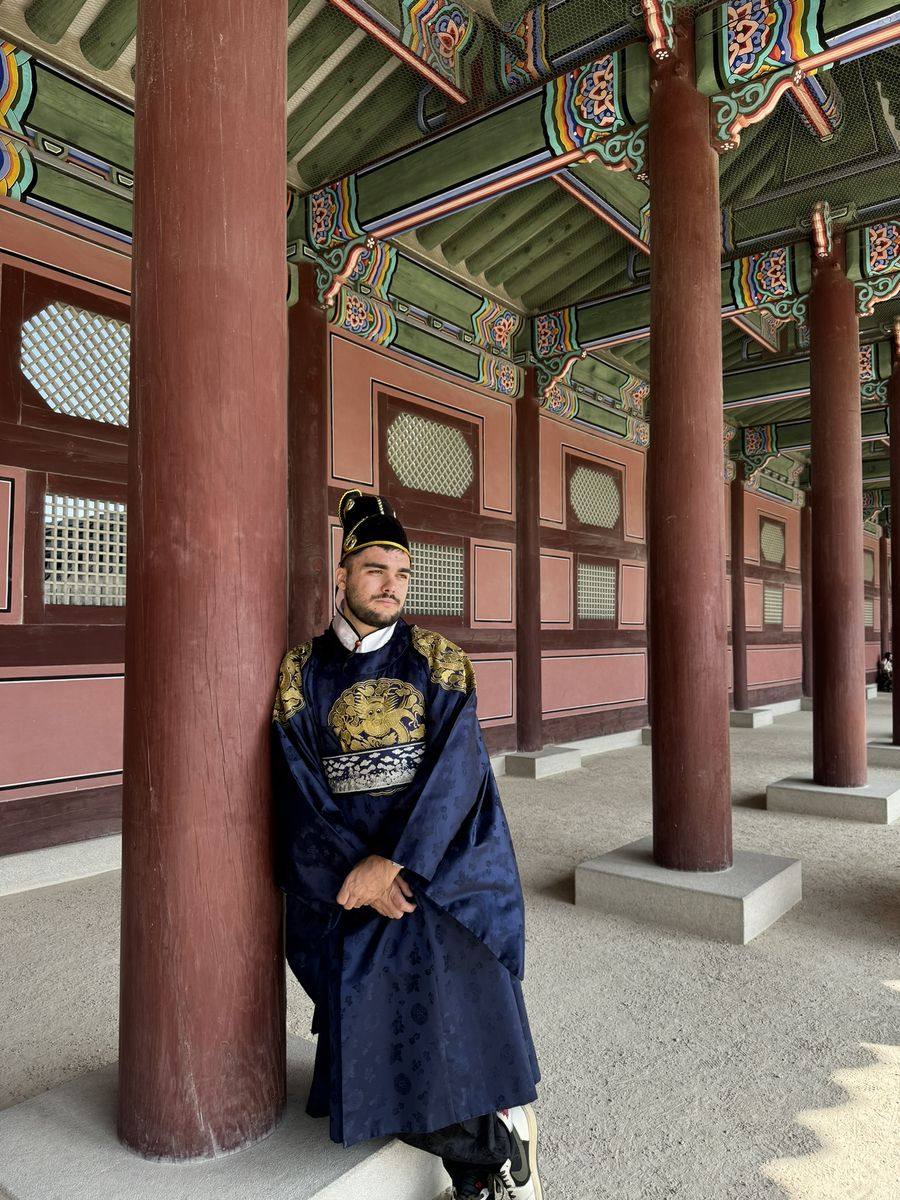 Moi en hanbok bleu nuit brodé d'un dragon doré, coiffé d'un chapeau noir, adossé à une colonne rouge sous une galerie aux poutres peintes de Gyeongbokgung