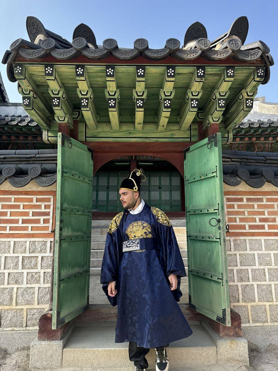 Moi en hanbok bleu nuit et chapeau noir à liseré doré, debout entre les deux battants verts d'une porte de Gyeongbokgung, sous un toit de tuiles grises