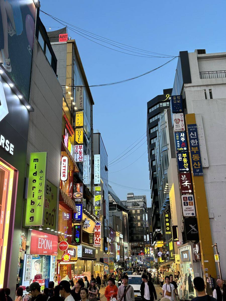 La rue commerçante de Myeongdong à la tombée de la nuit : enseignes lumineuses empilées sur les façades, foule dense sur la chaussée piétonne