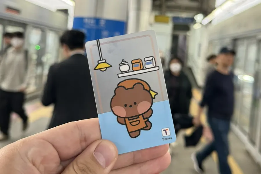 Ma carte T-money illustrée d'un petit ours en tablier, tenue à la main sur un quai de métro aérien de Séoul, des voyageurs flous en arrière-plan