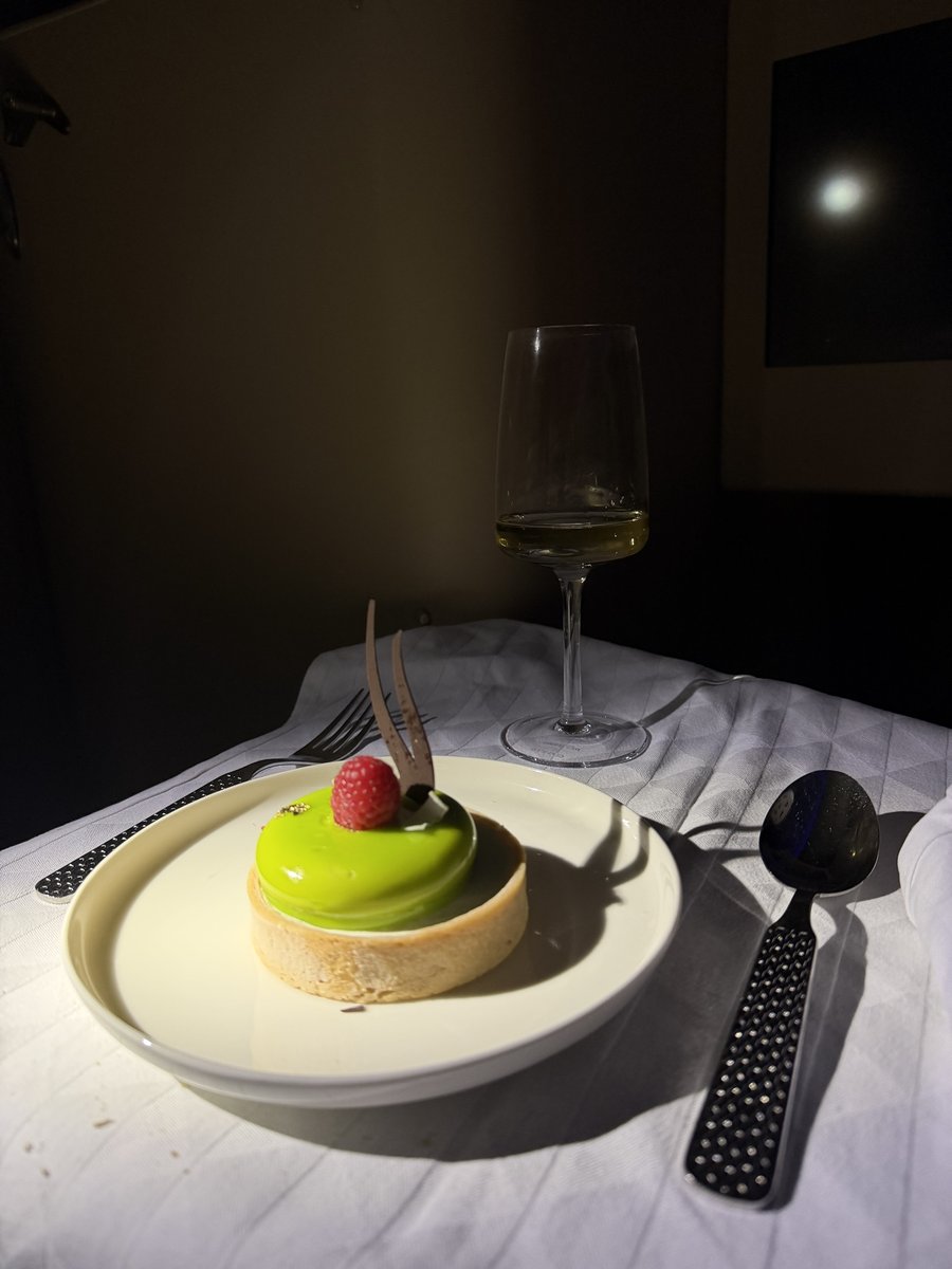 Tartelette au glaçage vert brillant surmontée d'une framboise et d'un éclat de chocolat, servie sur assiette blanche avec un verre de vin blanc dans la cabine assombrie du vol de nuit vers Bali