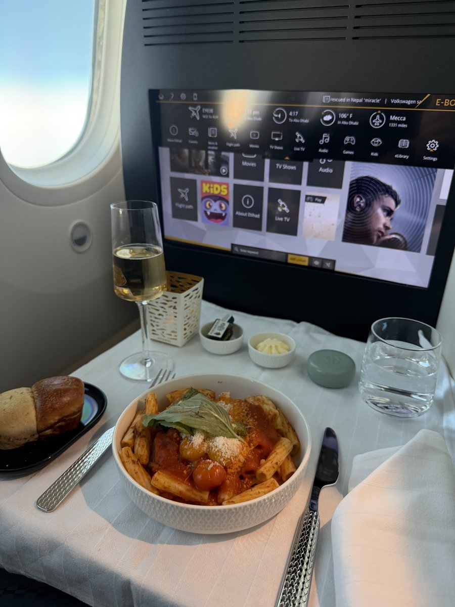 Rigatoni à la sauce tomate, tomates cerises, basilic et parmesan servis en bol sur nappe blanche en business class Etihad, avec pain, coupe de champagne et écran E-BOX affichant le vol Nice – Abu Dhabi