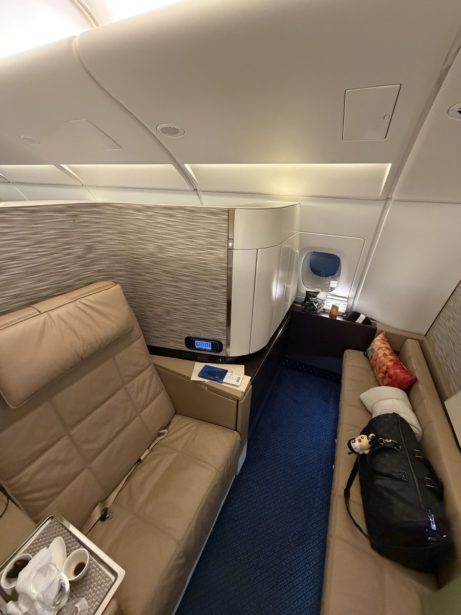 Vue d'ensemble d'un First Apartment de l'A380 Etihad : fauteuil en cuir beige à gauche, banquette convertible à droite, couloir privatif au sol bleu, minibar et coiffeuse au fond près du hublot