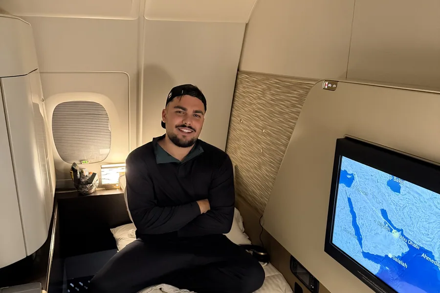 Monsieur Voyage assis en souriant sur le lit fait de son First Apartment Etihad, hublot à gauche et carte du vol vers Abu Dhabi à l'écran