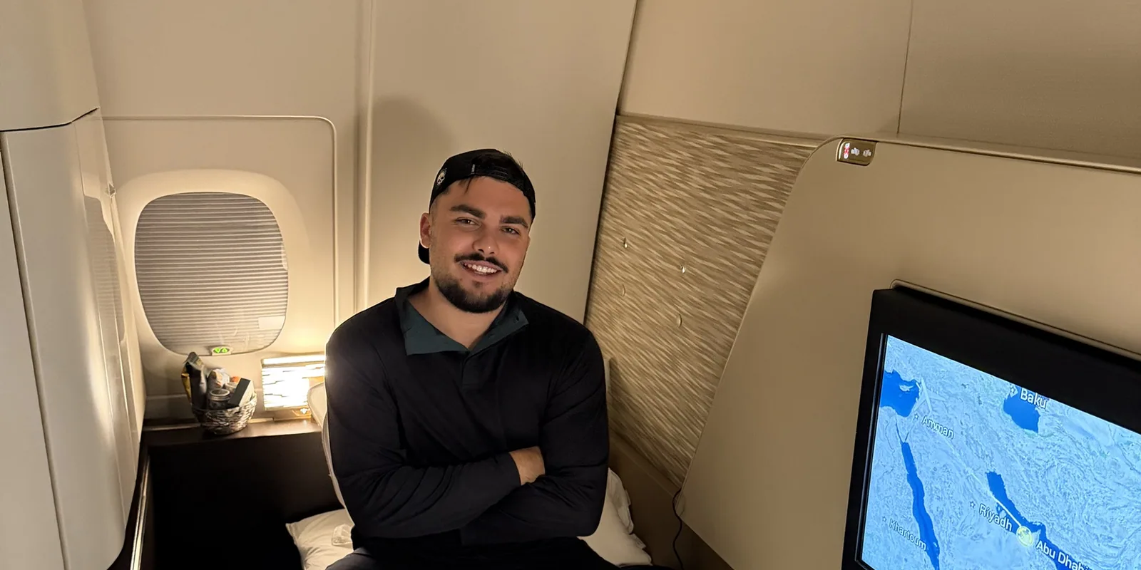 Monsieur Voyage assis en souriant sur le lit fait de son First Apartment Etihad, hublot à gauche et carte du vol vers Abu Dhabi à l'écran