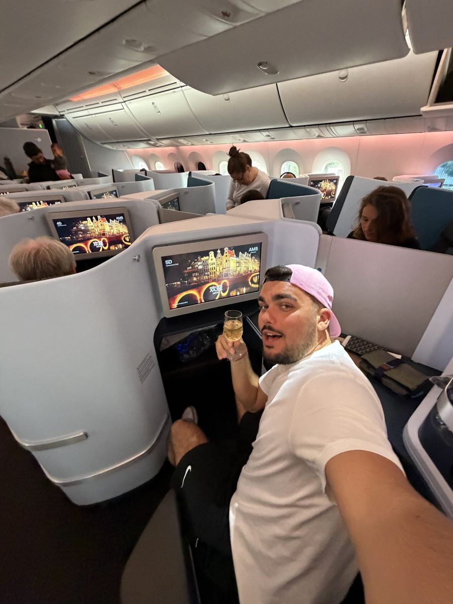Passager en classe affaires KLM, coupe de champagne à la main, écrans de bord affichant un message de bienvenue à Amsterdam