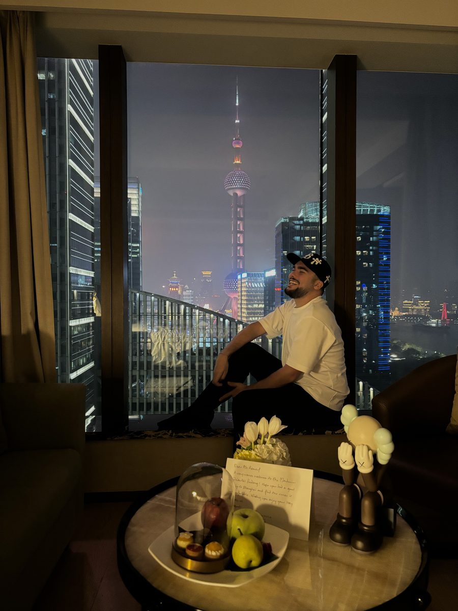 Monsieur Voyage assis devant la baie vitrée de sa chambre au Mandarin Oriental Pudong avec la tour de la Perle de l'Orient illuminée la nuit