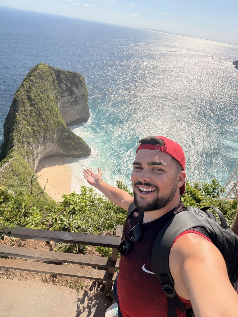 Moi en casquette rouge et sac à dos au belvédère de Kelingking, la falaise en forme de T-Rex et la plage en contrebas derrière moi