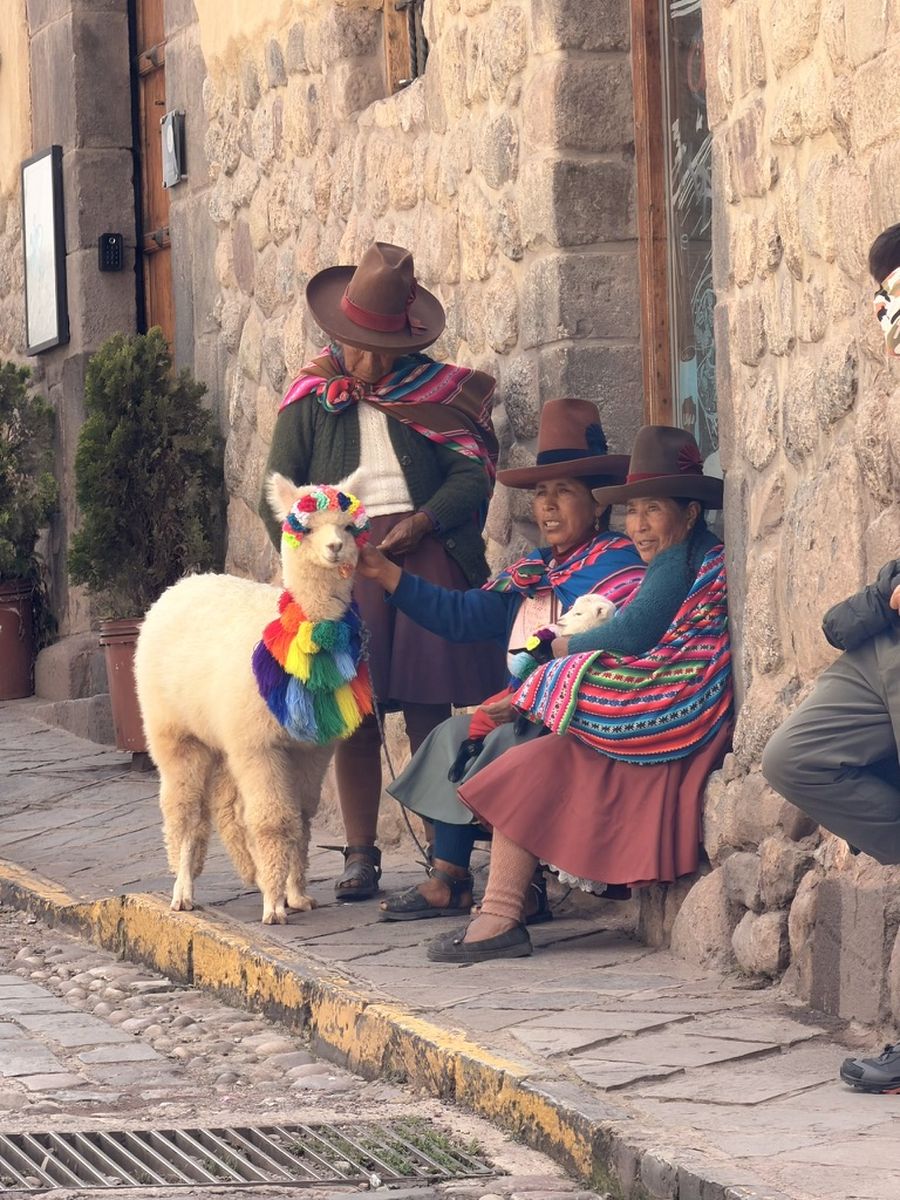 Un homme et deux femmes en tenue andine dans une ruelle pavée de Cuzco, avec un alpaga au collier multicolore