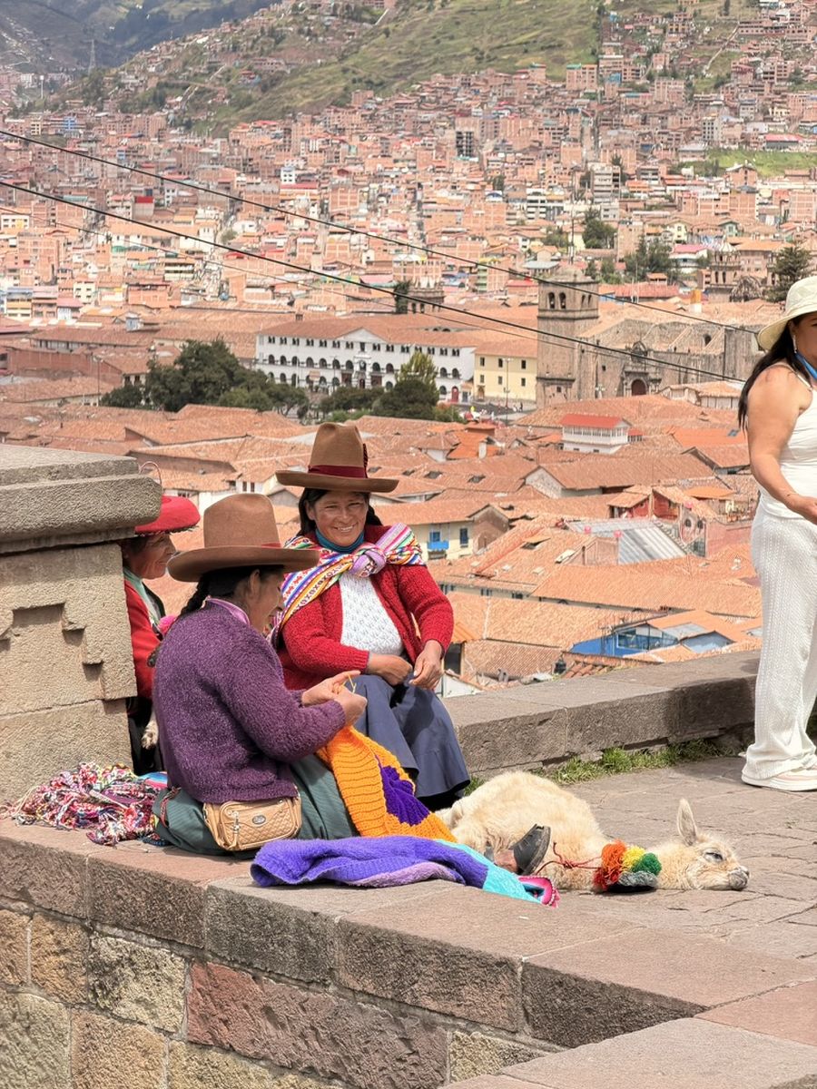 Deux femmes en tenue traditionnelle tricotent assises sur un muret, un alpaga couché à côté d'elles, les toits de tuiles de Cuzco en contrebas