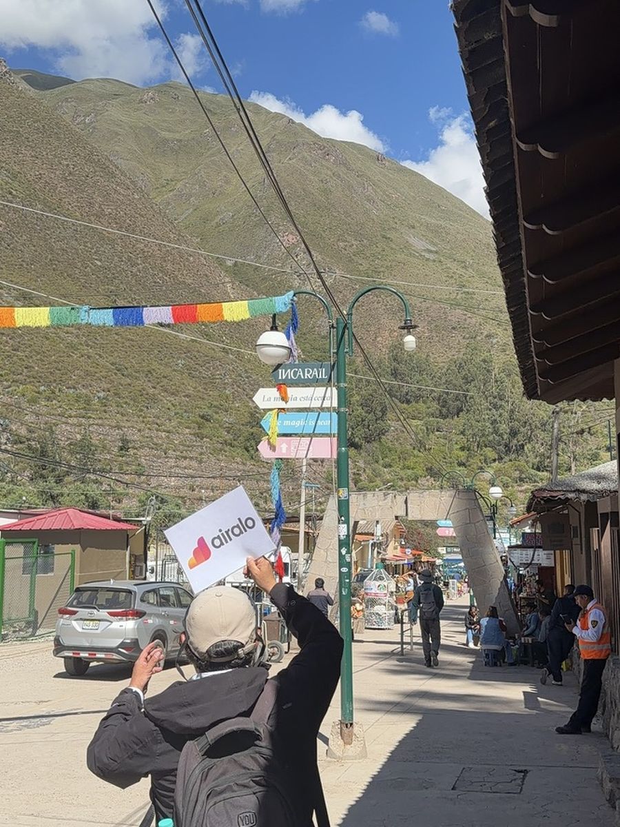 Ma main levant un panneau Airalo devant les panneaux de la gare d'Ollantaytambo, montagne andine en arrière-plan