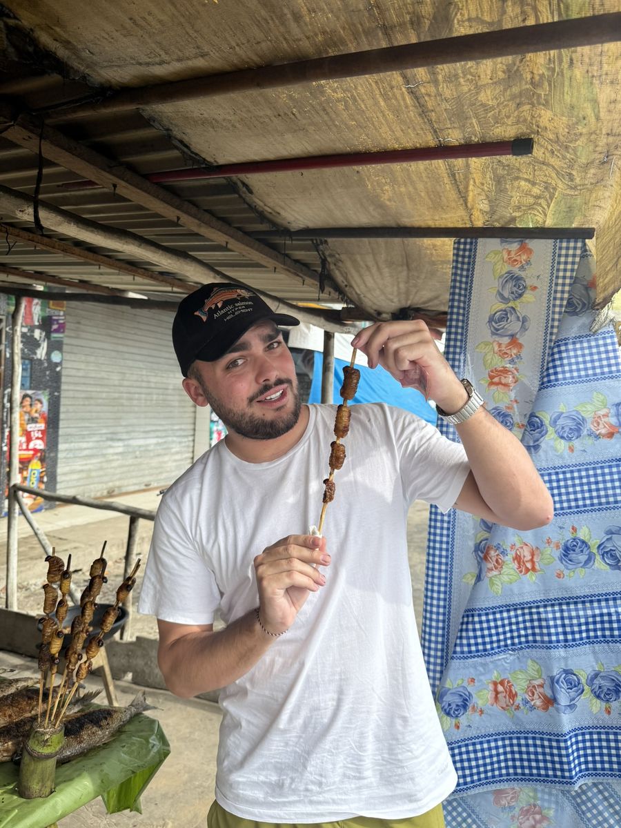 Moi tenant une brochette de larves de suri grillées devant un étal de marché à Iquitos, d'autres brochettes et des poissons grillés au premier plan