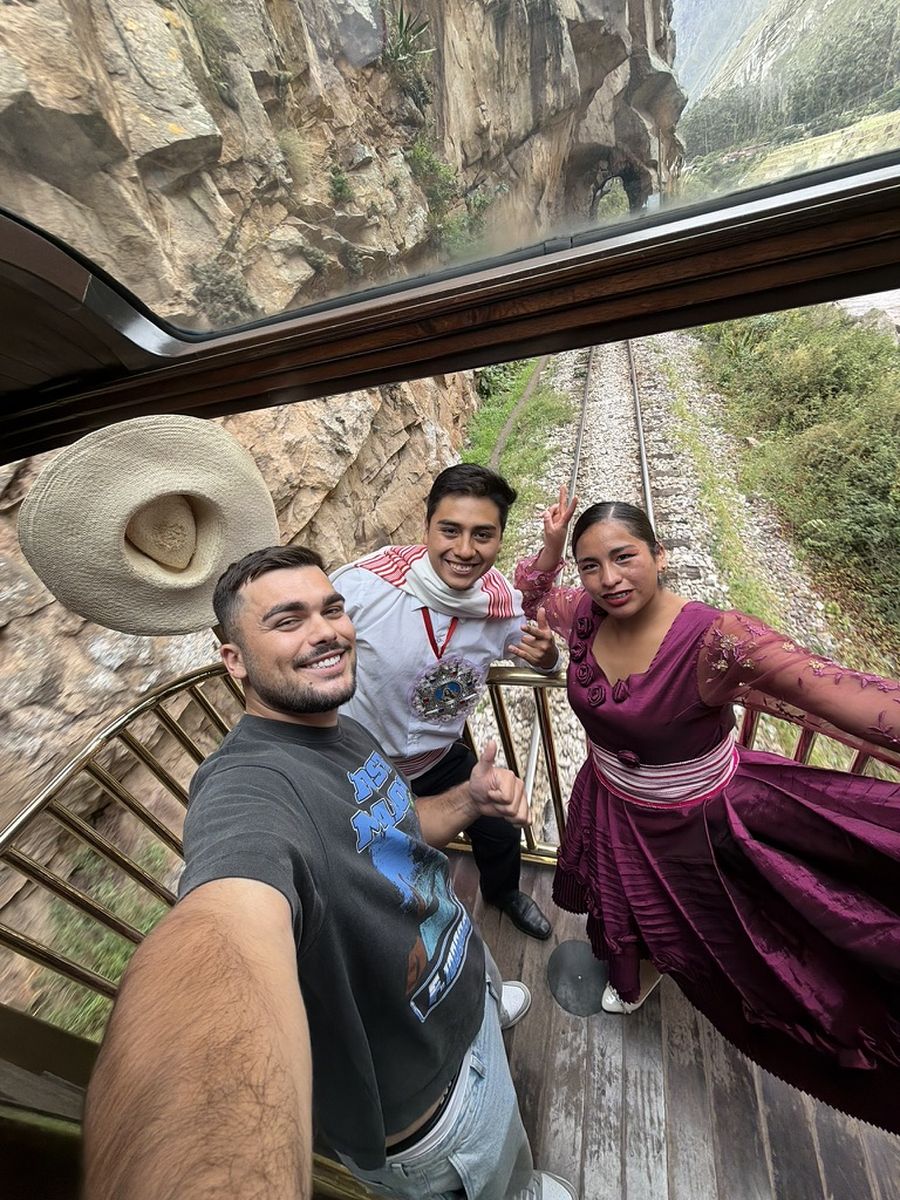 Selfie dans le wagon panoramique du Vistadome avec un membre d'équipage et une danseuse en tenue traditionnelle violette, la voie ferrée visible par la vitre du toit